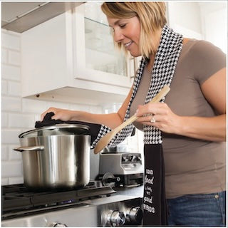 Woman cooking in a kitchen using the kitchen boa towel wrapped around her neck to take the lid off a pot
