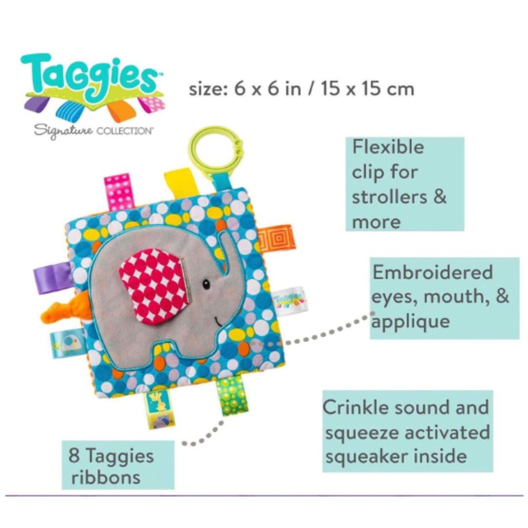 A baby activity toy labeled Taggies Signature Collection, featuring a blue patterned fabric square with a gray elephant applique, colorful elastic tabs, and attached ribbon tags. Text callouts describe features such as a flexible clip for strollers, embroidered eyes and mouth, crinkle sound and squeaker inside, and 8 Taggies ribbons.