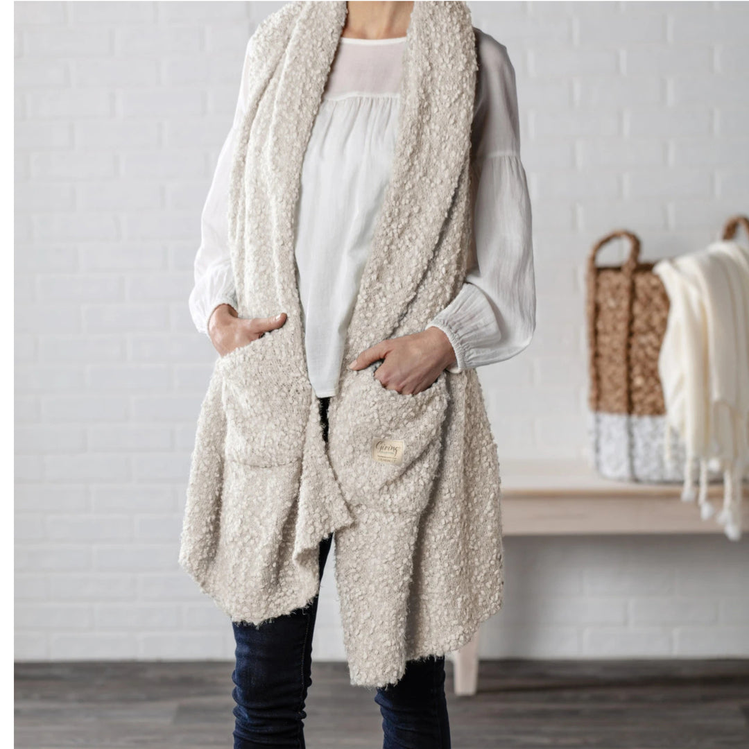 A person wearing a long, open-front sherpa vest shawl with 2 pocket in a warm off-white or cream color