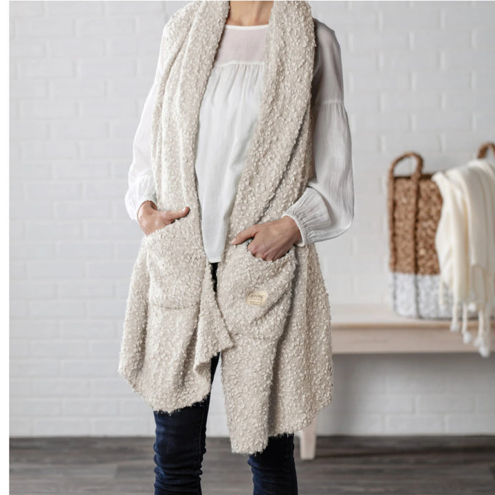 A person wearing a long, open-front sherpa vest shawl with 2 pocket in a warm off-white or cream color