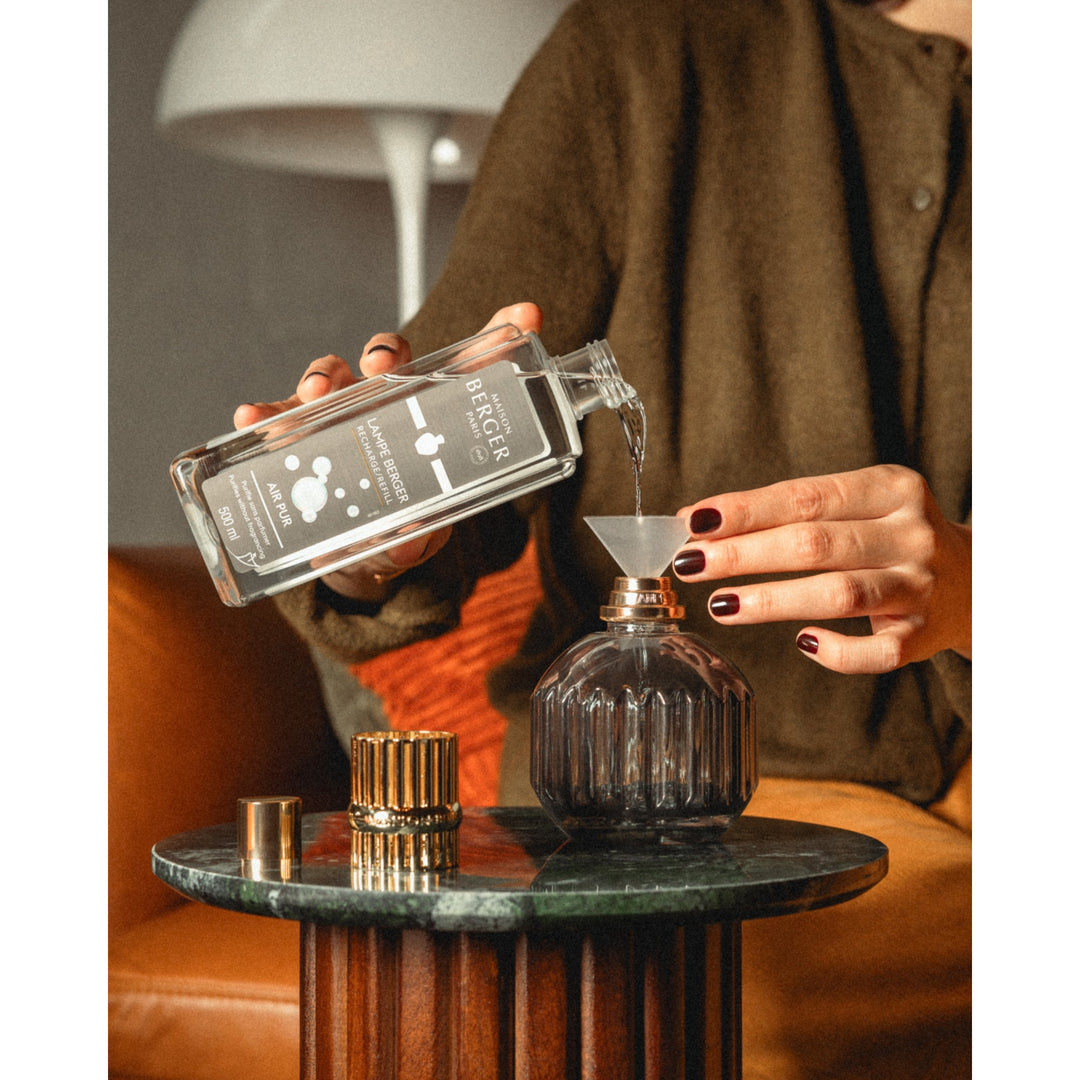 A person pouring fragrance through a funnel into a smoke-grey fluted Lampe Berger Cannelo lamp, with its gold-coloured top beside it.