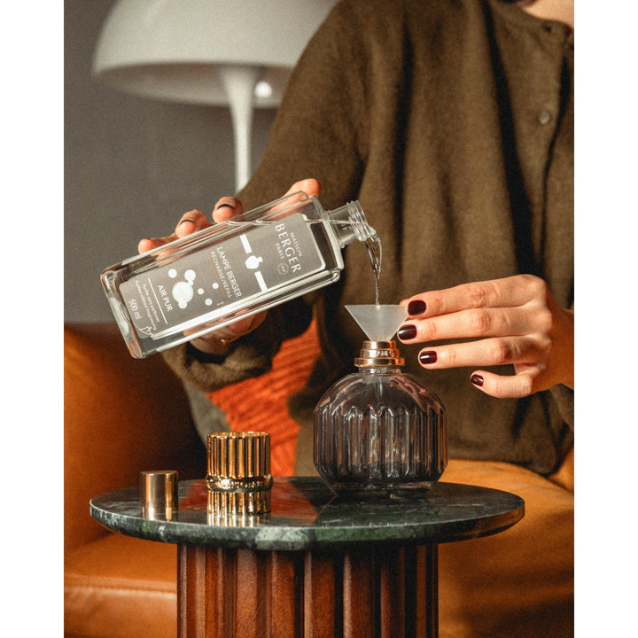 A person pouring fragrance through a funnel into a smoke-grey fluted Lampe Berger Cannelo lamp, with its gold-coloured top beside it.