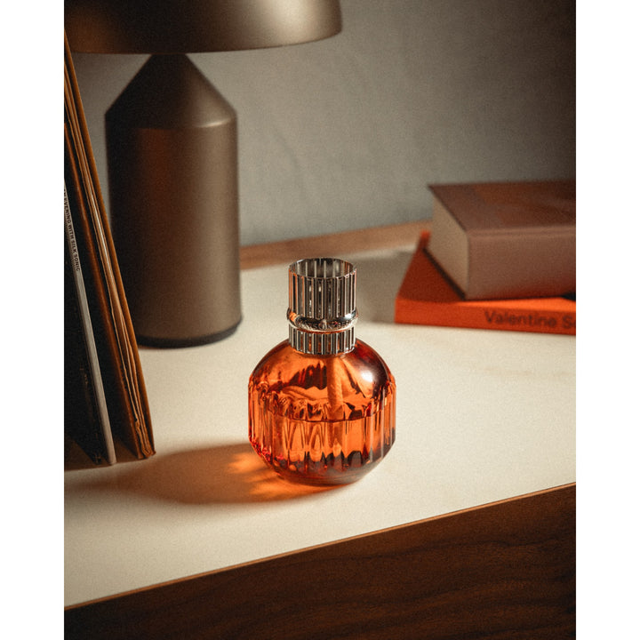 Lampe Berger Cannelo Paprika lamp in warm amber-orange fluted glass with a silver-coloured slotted diffuser top, displayed beside books and a table lamp.
