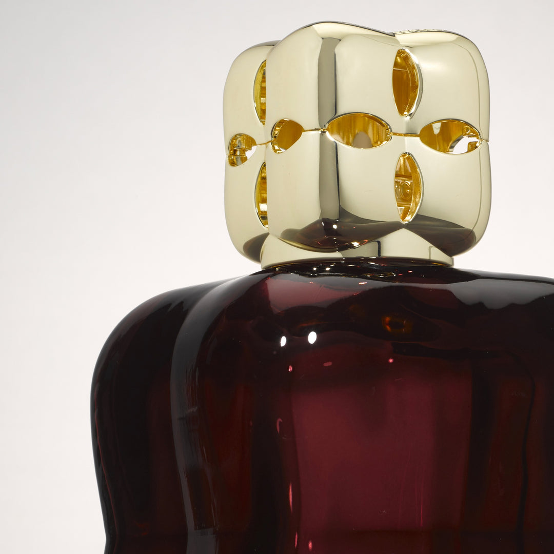 Close-up of the gold-coloured diffuser cap with oval cut-outs and deep plum glass with softly undulating sides of the Lampe Berger Cosy Plum home fragrance lamp.
