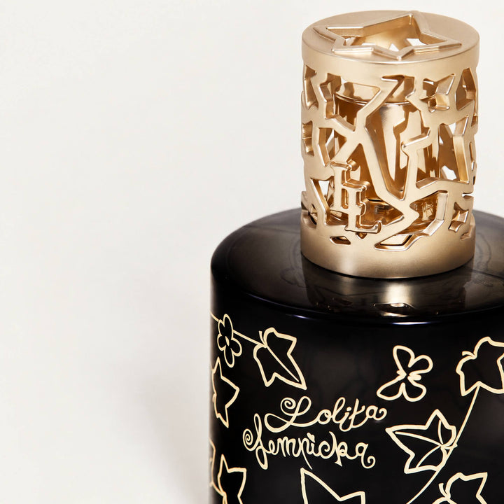 A close-up of a black Lolita Lempicka  bottle with gold floral art and the script brand name 'Lolita Lempicka' on the front, topped with a gold cylindrical cap featuring an intricate cutout pattern.