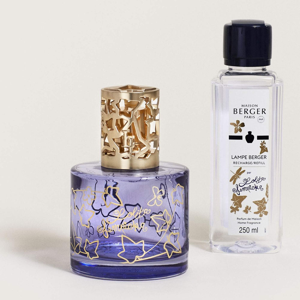 Lampe Berger Lolita Lempicka home fragrance lamp with a cylindrical violet-blue glass body, gold ivy-leaf outlines and Lolita Lempicka script, topped by a satin-gold ivy-cutout diffuser cap, displayed beside the matching 250 ml Lolita Lempicka fragrance refill bottle.
