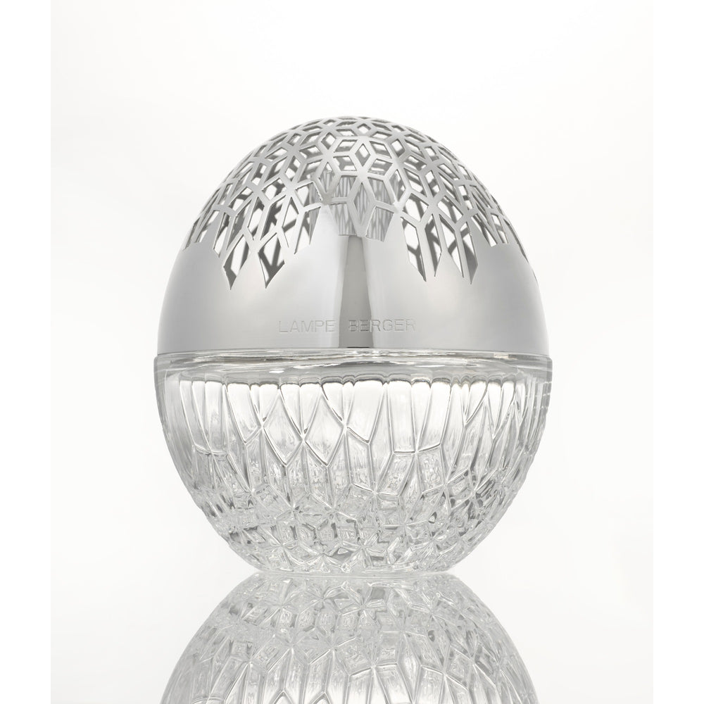 Luka lampe berger A clear cut-glass egg-shaped  sphere with a polished silver lid featuring a geometric laser-cut pattern, shown on a reflective white surface.