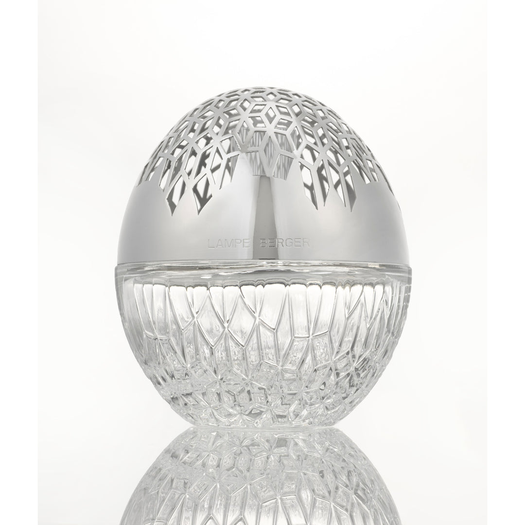 Luka lampe berger A clear cut-glass egg-shaped  sphere with a polished silver lid featuring a geometric laser-cut pattern, shown on a reflective white surface.