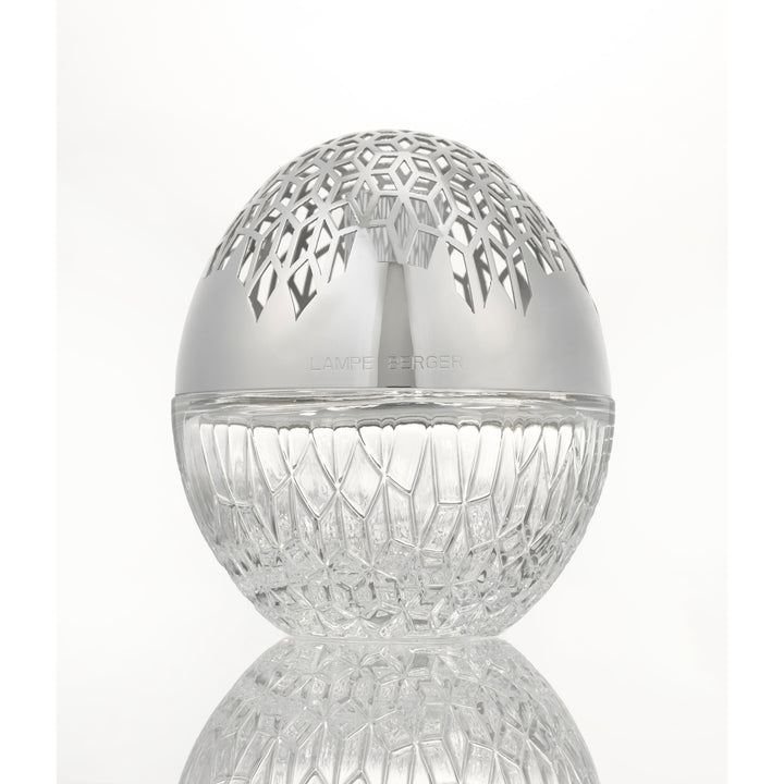 Luka lampe berger A clear cut-glass egg-shaped  sphere with a polished silver lid featuring a geometric laser-cut pattern, shown on a reflective white surface.